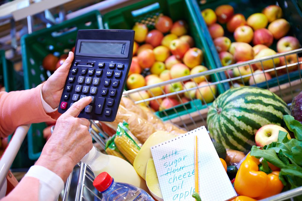 Senior woman holding up calculator in front of full shopping cart.