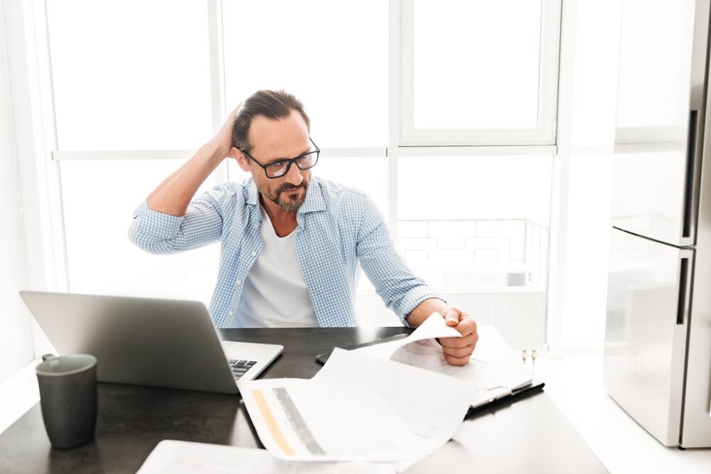 Confused mature man working with documents while sitting with laptop computer at the table at home