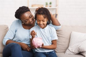 African Mother And Daughter Putting Coins Into Piggy Bank At Home. Free space