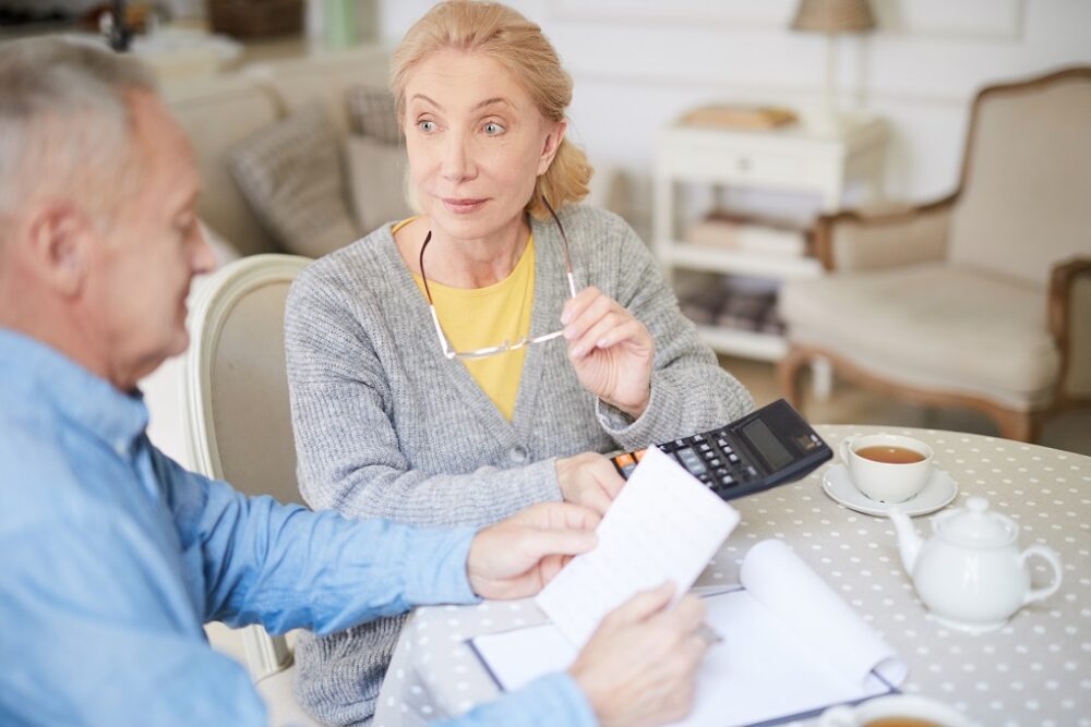 Senior woman with calculator talking to her husband while making calculations in the kitchen