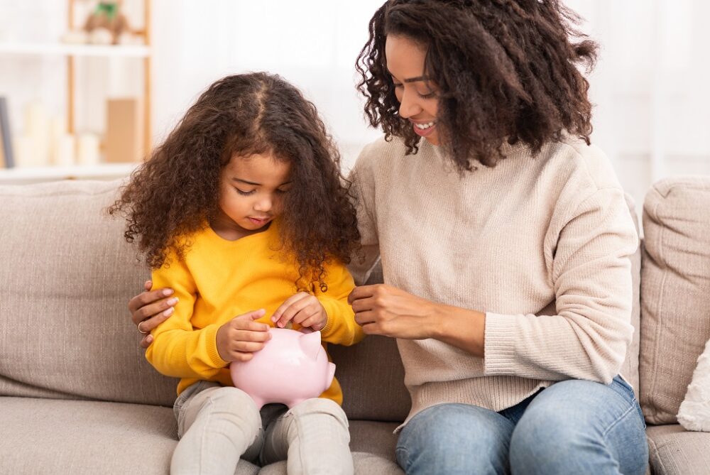 Personal Savings. Little African Girl Putting Money To Piggybank Sitting With Mother On Couch Indoor