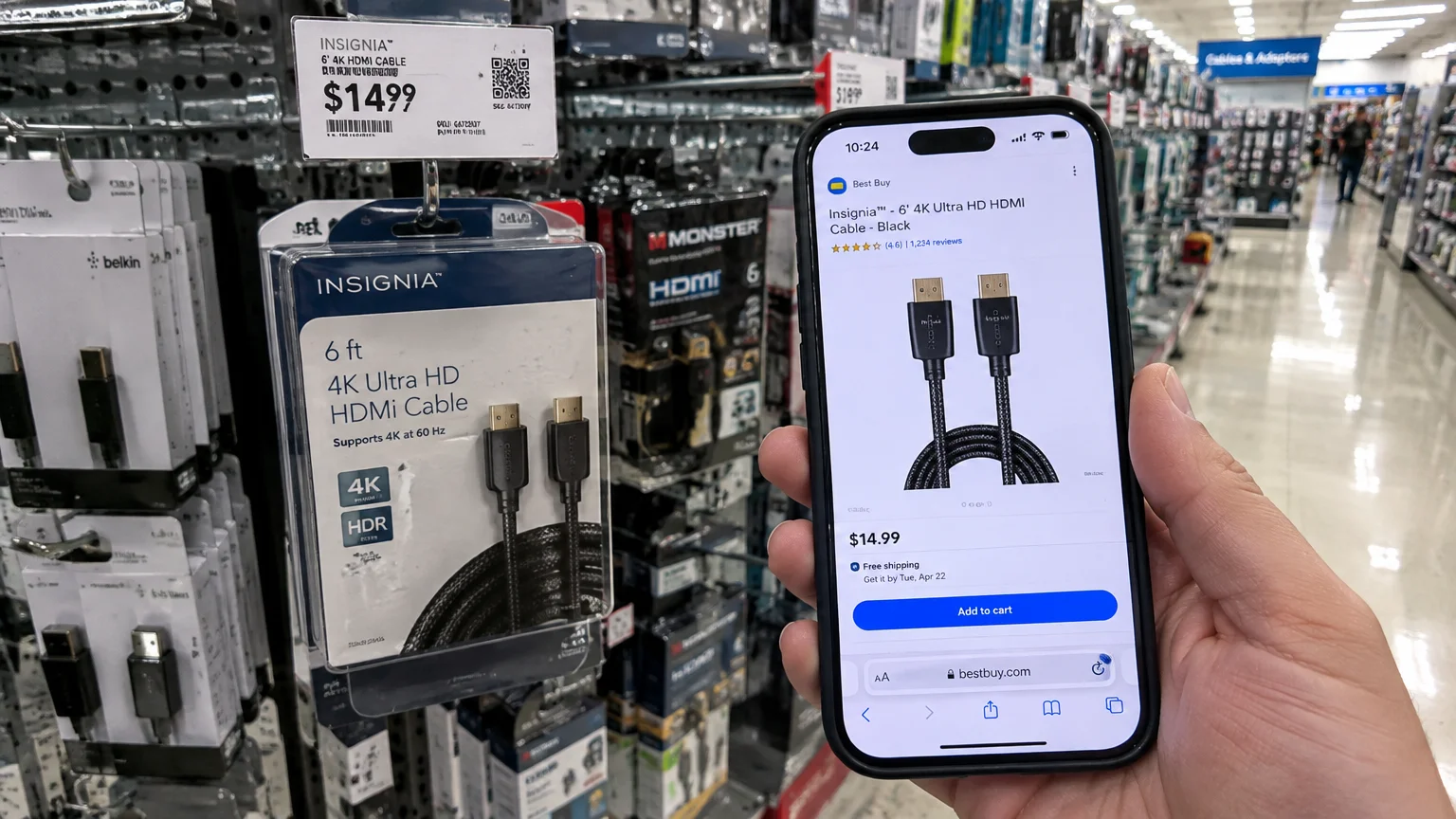 Hand holding a phone displaying a Best Buy webpage next to a boxed Insignia HDMI cable on a store shelf.