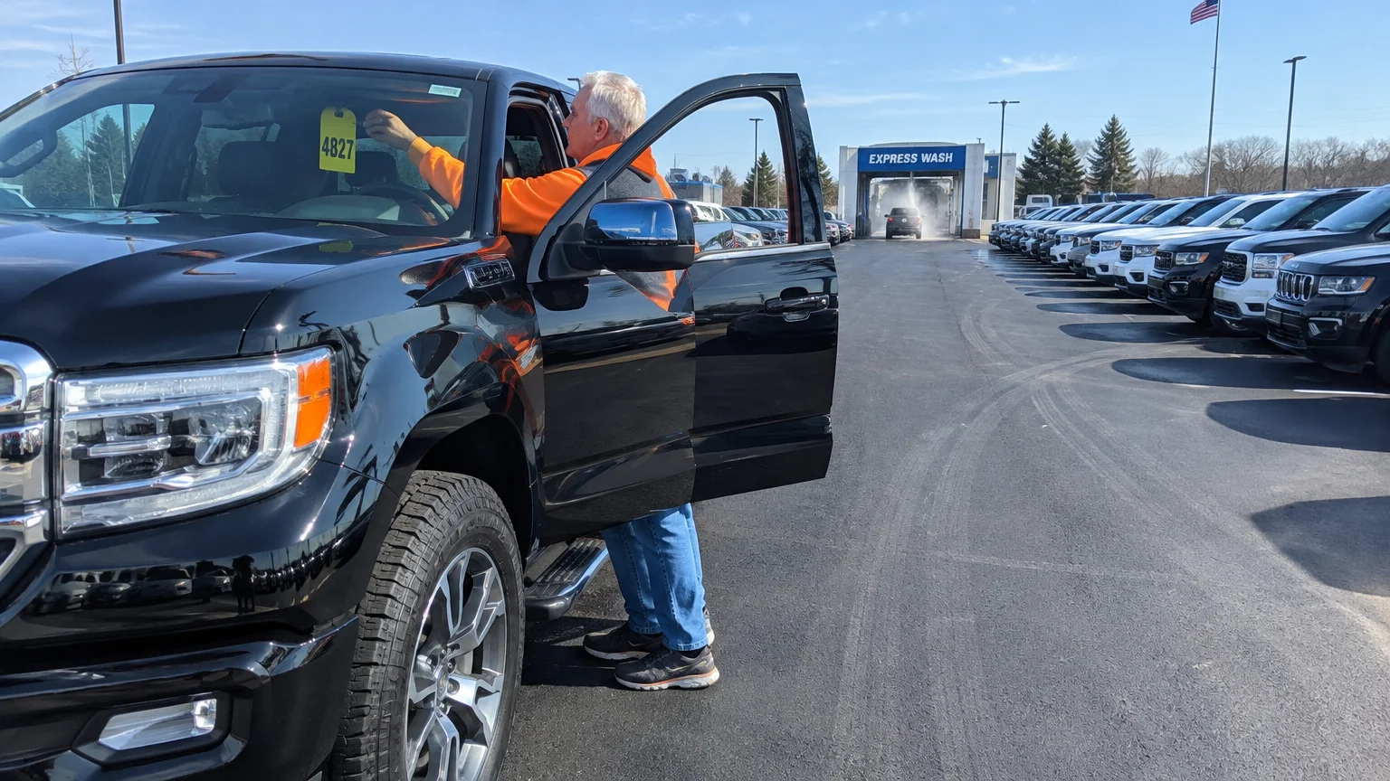 A man placing a numbered yellow tag on the rearview mirror of a black pickup truck on a dealership lot.