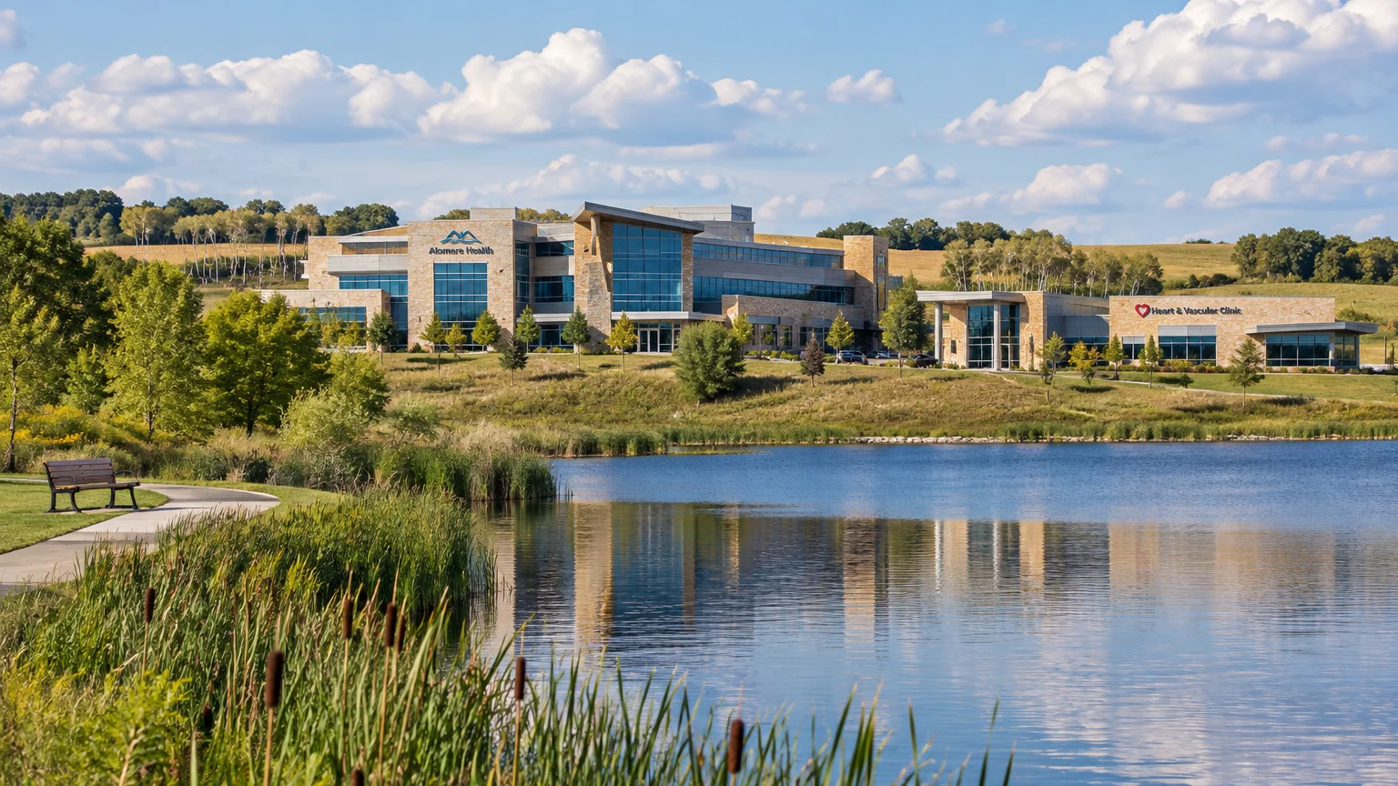 Modern stone and glass Alomere Health building and clinic reflecting across a pond bordered by reeds and a walking path.